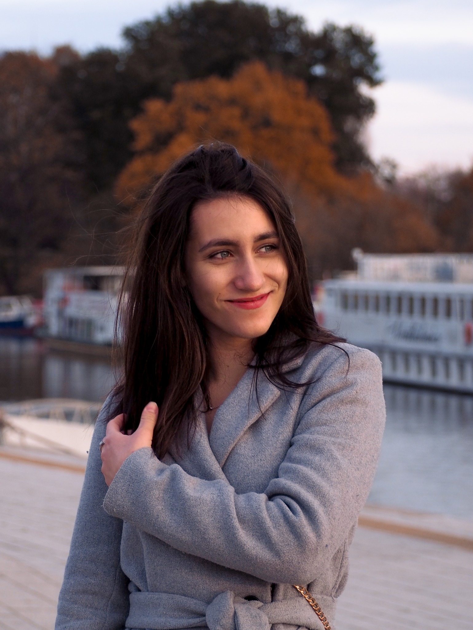 Portret uśmiechniętej kobiety w szarym płaszczu na tle jesiennego parku i rzeki z łodziami.