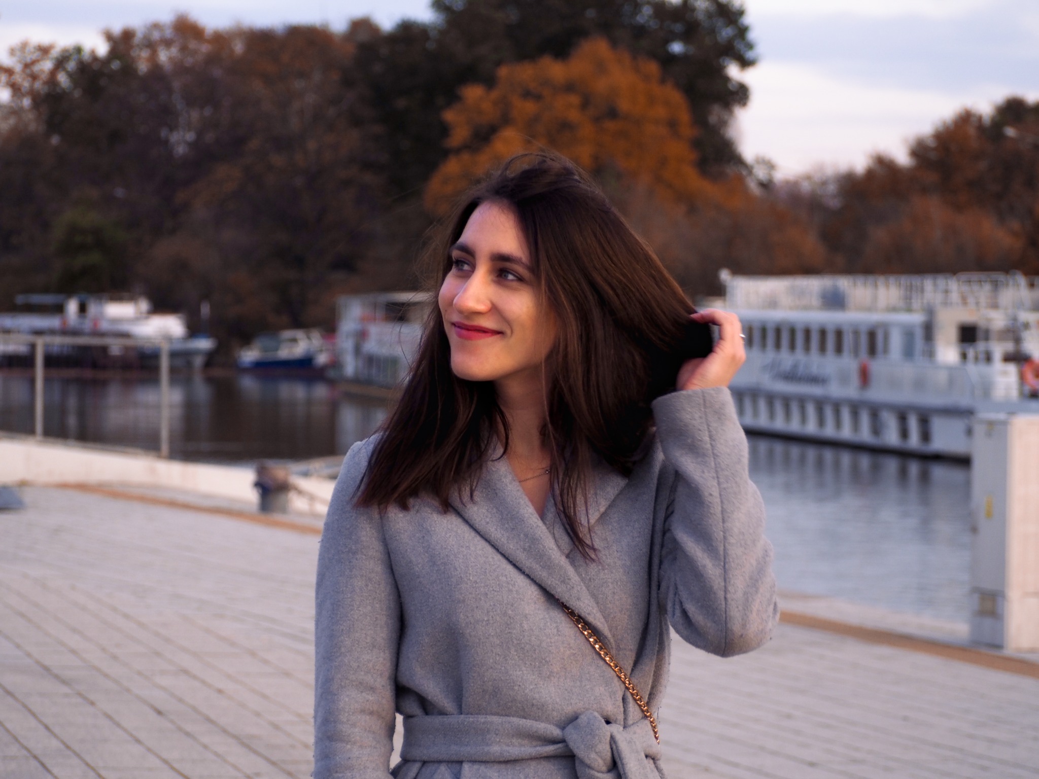 Uśmiechnięta kobieta w szarym płaszczu, z torebką na łańcuszku, poprawia włosy na tle jesiennego krajobrazu z wodą i budynkami.