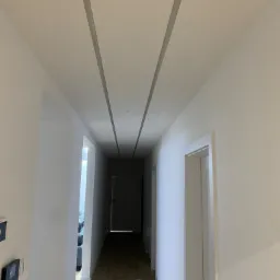 Korytarz w jasnych kolorach z panelami na suficie i widocznym wejściem do pokoju. Na podłodze leżą białe materiały zabezpieczające.