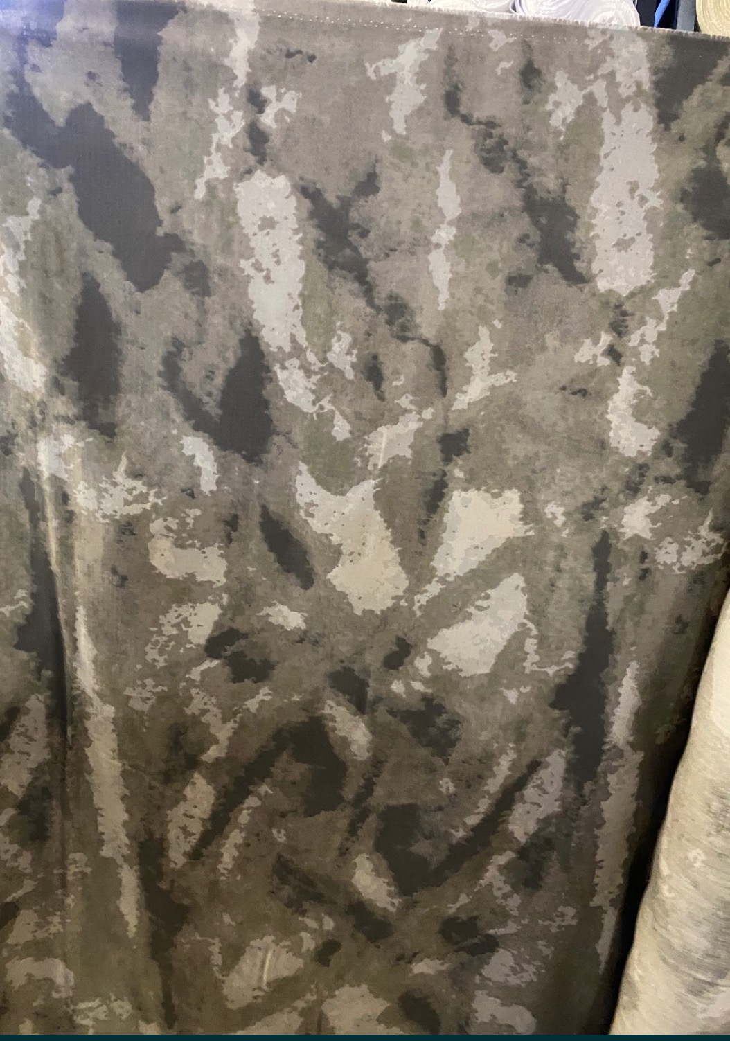 Bliskie ujęcie tkaniny w odcieniach szarości, zieleni i bieli, przypominającej wzór kamuflażu, z widocznymi fałdami i teksturą materiału.