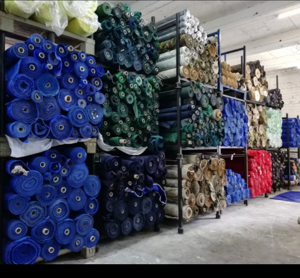 Magazyn z półkami wypełnionymi rolkami tkanin w różnych kolorach, od błękitu po zieleń i beż, ułożonych warstwami na metalowych regałach.