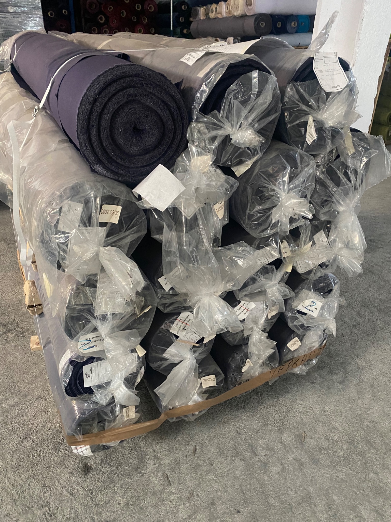 Paleta z belami ciemnej tkaniny, owinięte folią i zabezpieczone etykietami, składowane w magazynie.