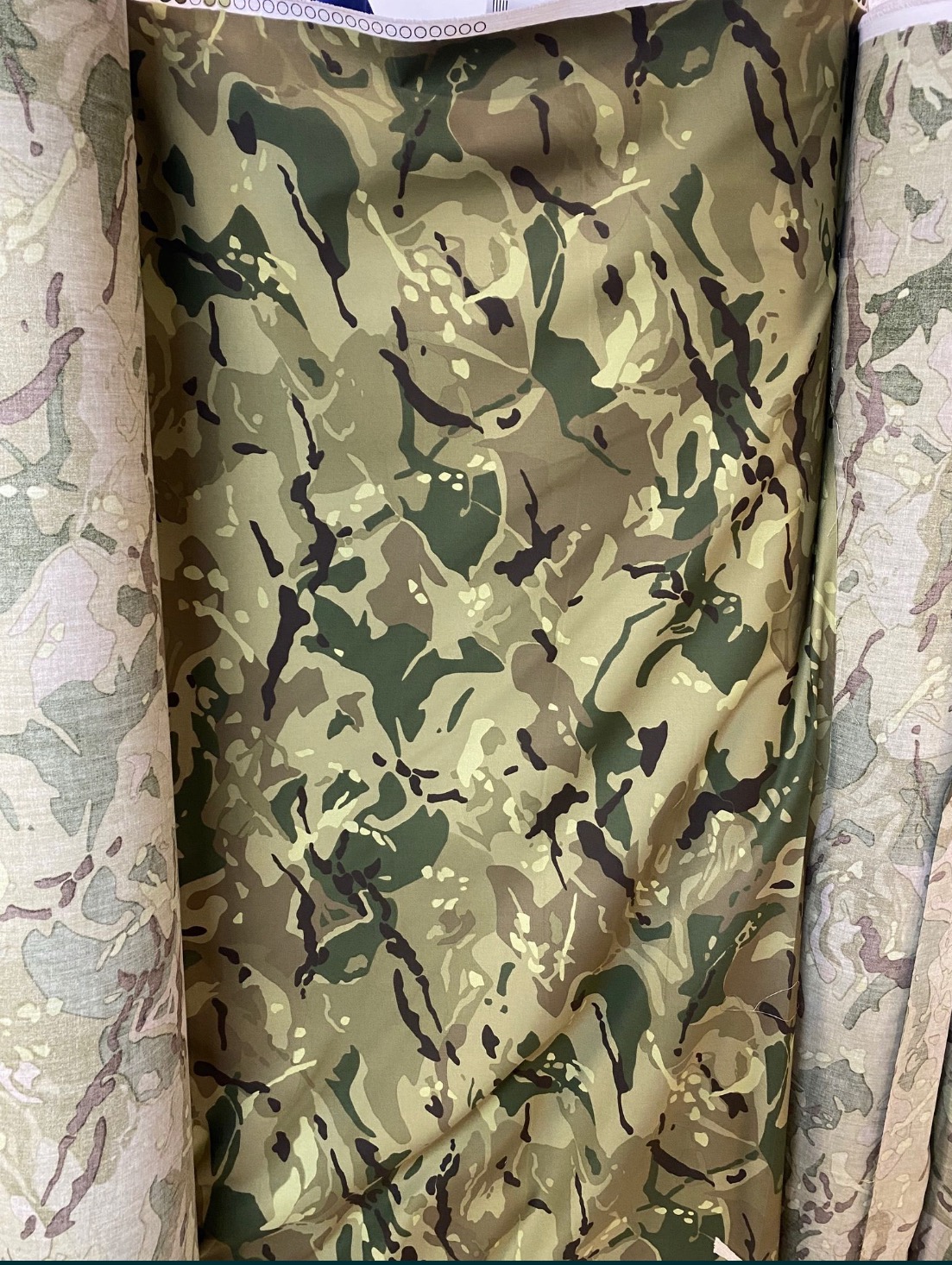 Rolki tkanin z kamuflażem w odcieniach zieleni, brązu i beżu, widoczne z bliska w magazynie.