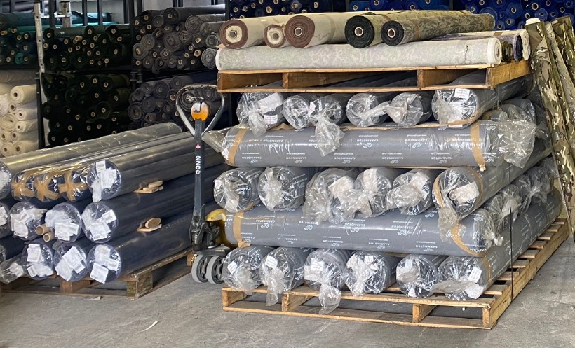 Magazyn tkanin: palety z rolkami materiałów w foliowych opakowaniach, obok wózek paletowy i regały z tkaninami w tle.