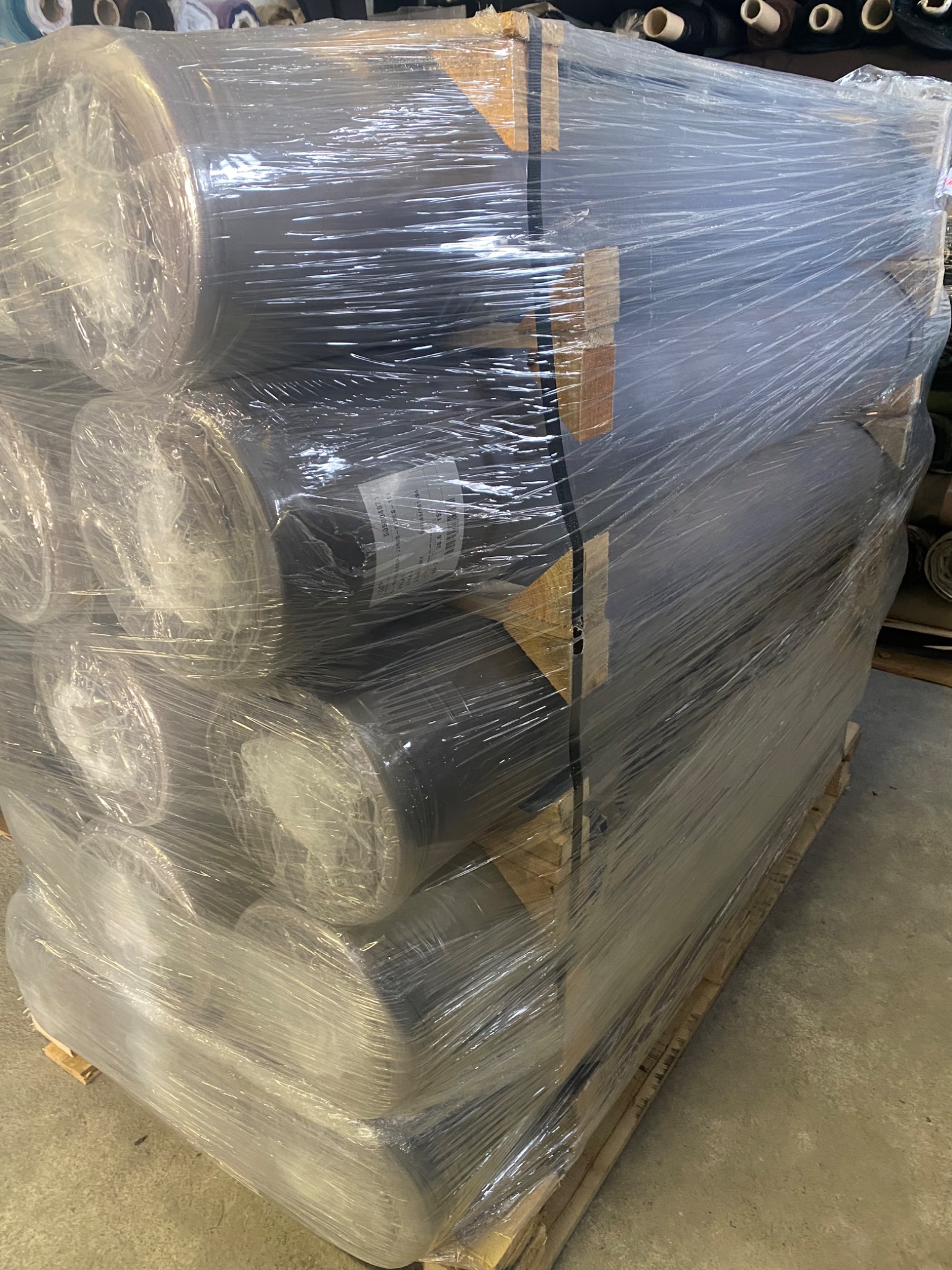 Paleta z rolkami ciemnej tkaniny, szczelnie owinięta folią, widoczne etykiety na niektórych rolkach.