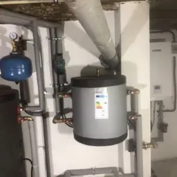 Szary zasobnik ciepłej wody użytkowej z etykietą energetyczną, podłączony do rur i armatury w kotłowni, z widoczną instalacją odprowadzającą spaliny pod sufitem.