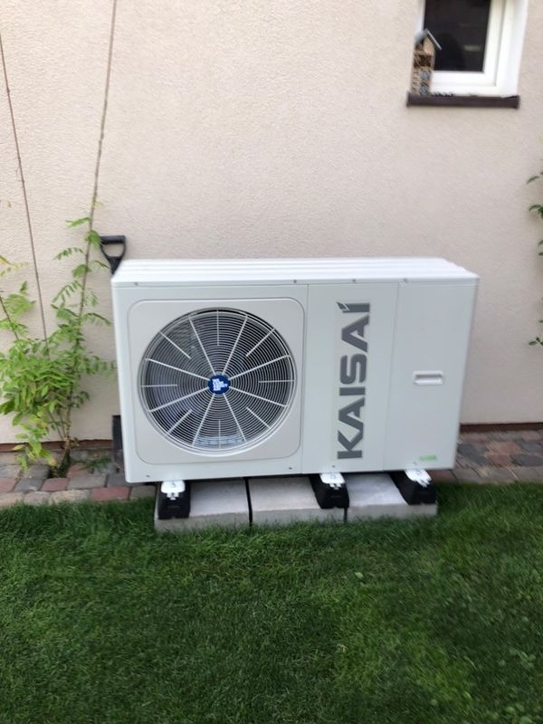 Biała zewnętrzna jednostka pompy ciepła Kaisai, umieszczona na betonowych bloczkach na trawniku przy ścianie budynku.