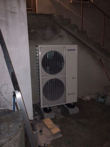 Biała, dwuczęściowa jednostka zewnętrzna klimatyzatora marki Samsung, umieszczona na betonowych bloczkach na nierównym podłożu, z widocznymi elementami konstrukcyjnymi budynku w tle.