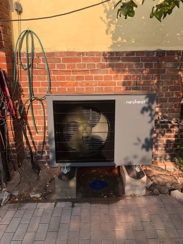 Szara pompa ciepła marki Nesheat zamontowana na betonowych blokach przy ceglanej ścianie budynku w Toruniu, widoczny fragment węża ogrodowego i narzędzi.
