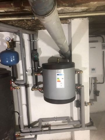 Szary zasobnik ciepłej wody użytkowej z etykietą energetyczną, podłączony do rur i armatury w kotłowni, z widoczną instalacją odprowadzającą spaliny pod sufitem.