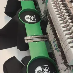 Wielogłowicowa maszyna hafciarska w trakcie pracy, haftuje biały herb na czarnych elementach odzieży, widoczne zielone tamborki i detale mechaniczne maszyny.