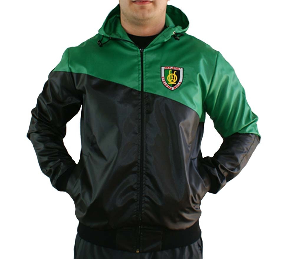 Mężczyzna w zielono-czarnej, ortalionowej kurtce z kapturem i naszywką herbu na piersi, stojący na białym tle.