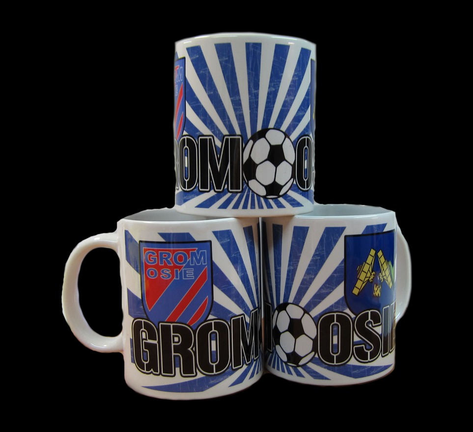 Trzy białe kubki z nadrukiem herbu, nazwy drużyny sportowej i piłki ułożone w piramidę na czarnym tle.