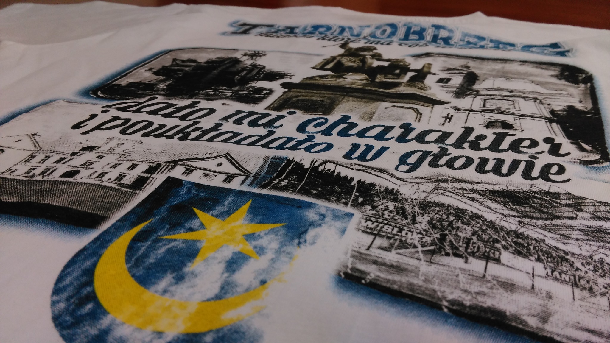 Biała koszulka z nadrukiem kolażu czarno-białych zdjęć i niebiesko-żółtego herbu, z napisem odnoszącym się do Tarnobrzega, wykonana w Grudziądzu.