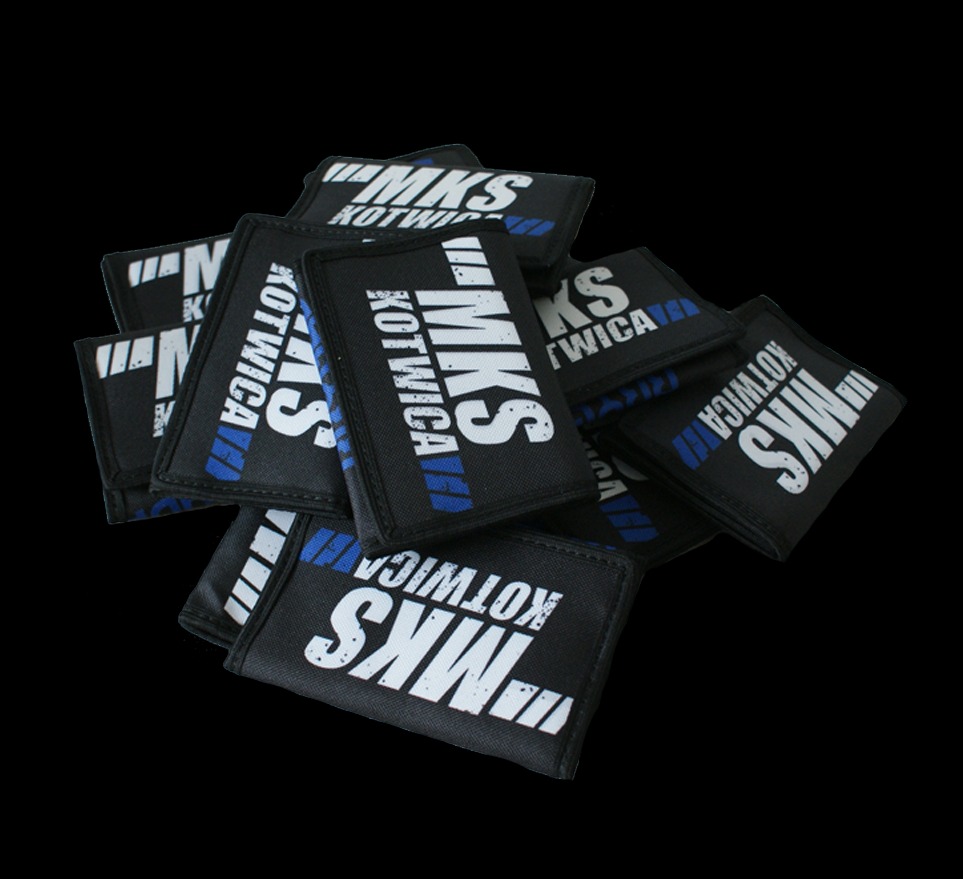 Stos czarnych, materiałowych portfelików z białym napisem 'MKS Kotwica' i niebieskim paskiem, ułożonych na czarnym tle.