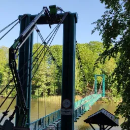 Zielony most wiszący dla pieszych i rowerzystów nad spokojną wodą, otoczony drzewami w słoneczny dzień.