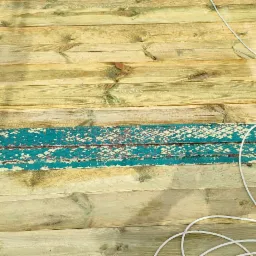 Pomost z desek pomalowanych na żółto-zielony kolor, otoczony turkusową metalową konstrukcją z widocznymi śladami korozji i odchodzącej farby, z leżącym obok białym kablem.