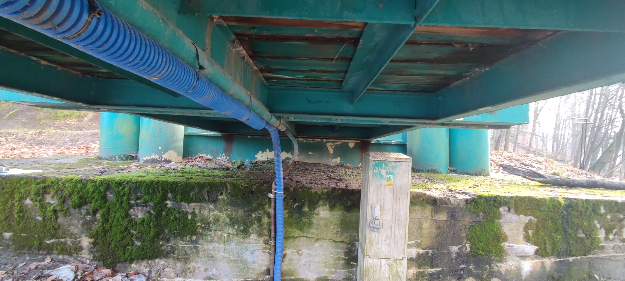 Spodnia część konstrukcji pomostu z widocznymi niebieskimi rurami ochronnymi na kable, zarośnięta mchem betonowa podstawa i słupki pomalowane na niebiesko-zielony kolor.