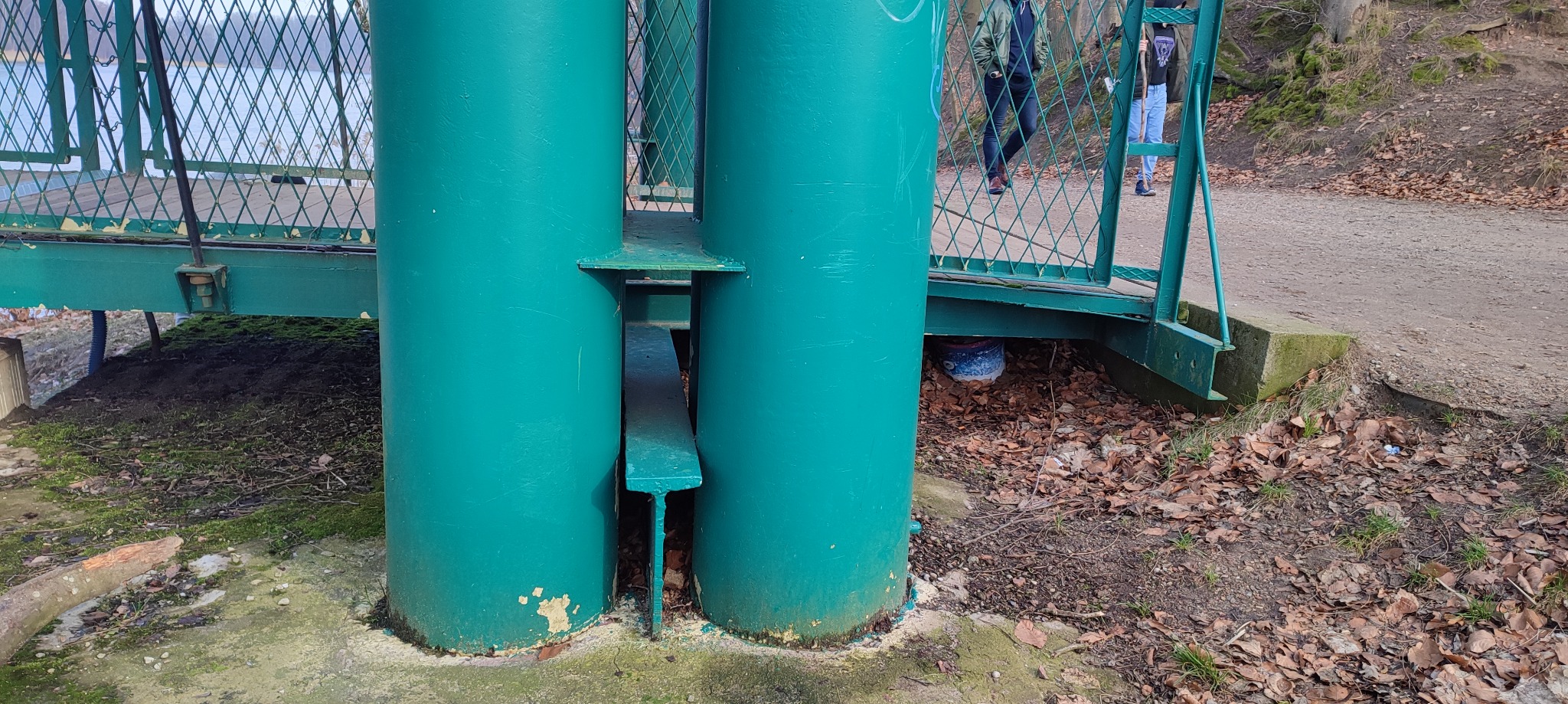 Zielona, metalowa konstrukcja mostu z widocznymi elementami spawania i śladami użytkowania, z widocznym fragmentem siatki zabezpieczającej i osobami w tle.