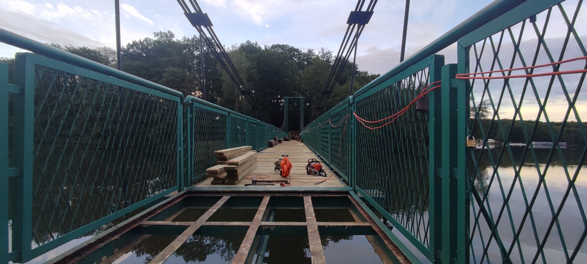 Remont kładki dla pieszych z zielonymi metalowymi barierkami, widoczne deski, piła łańcuchowa i dmuchawa do liści.