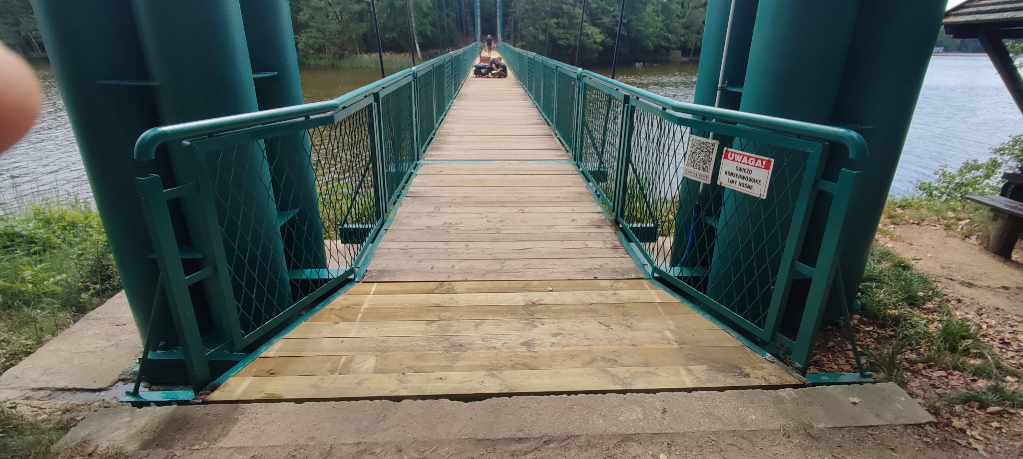 Zielony most wiszący z drewnianym pomostem nad jeziorem, tabliczka ostrzegawcza o konserwowanych linach nośnych, osoba siedząca na moście w oddali.