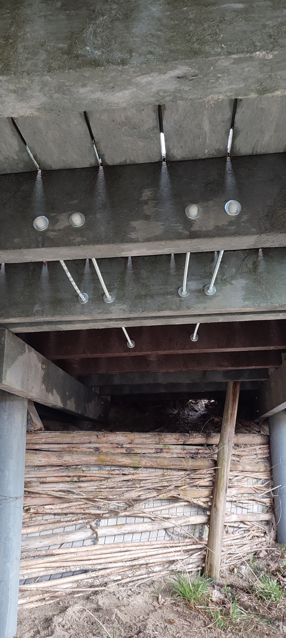 Widok od dołu na betonową konstrukcję z zamontowanymi punktowymi lampami i metalowymi prętami, z drewnianym wypełnieniem i rurą w tle.