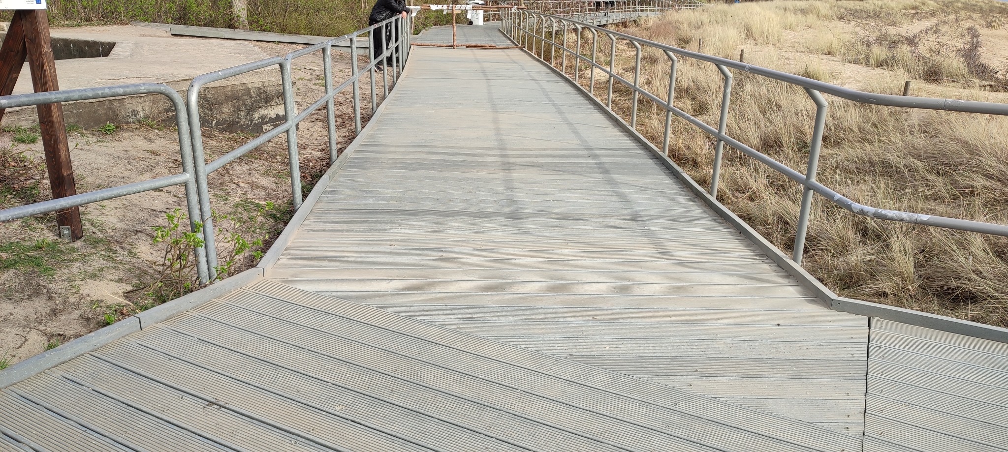 Długa, drewniana kładka z metalowymi barierkami prowadząca przez wydmy w kierunku plaży, widoczna osoba w oddali.