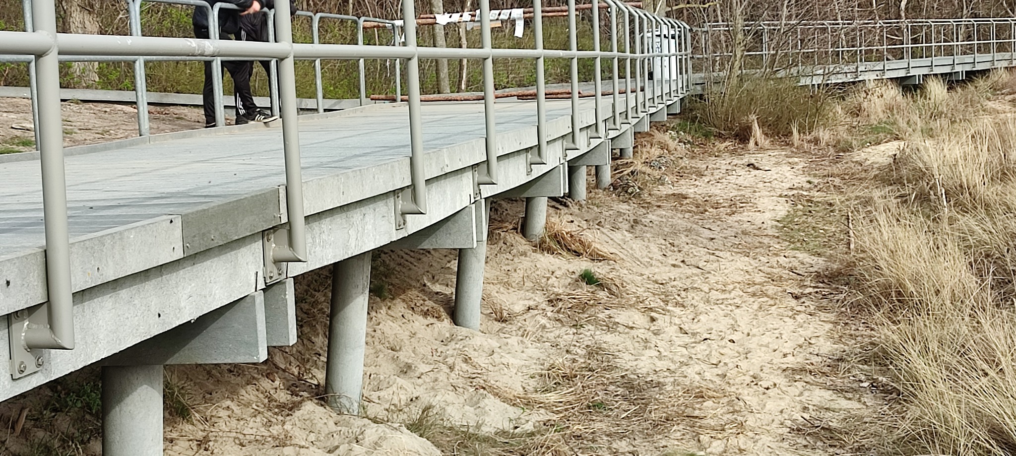 Długa, betonowa kładka z metalową barierką, wsparta na betonowych filarach, prowadząca przez piaszczysty teren z suchą trawą.