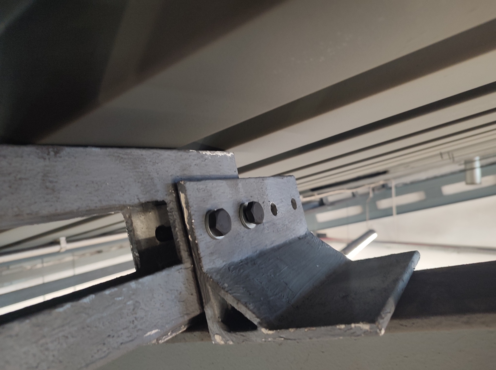 Szara, metalowa konstrukcja wsporcza z trzema śrubami mocującymi, widok z dołu, fragment sufitu z widocznymi elementami instalacji.