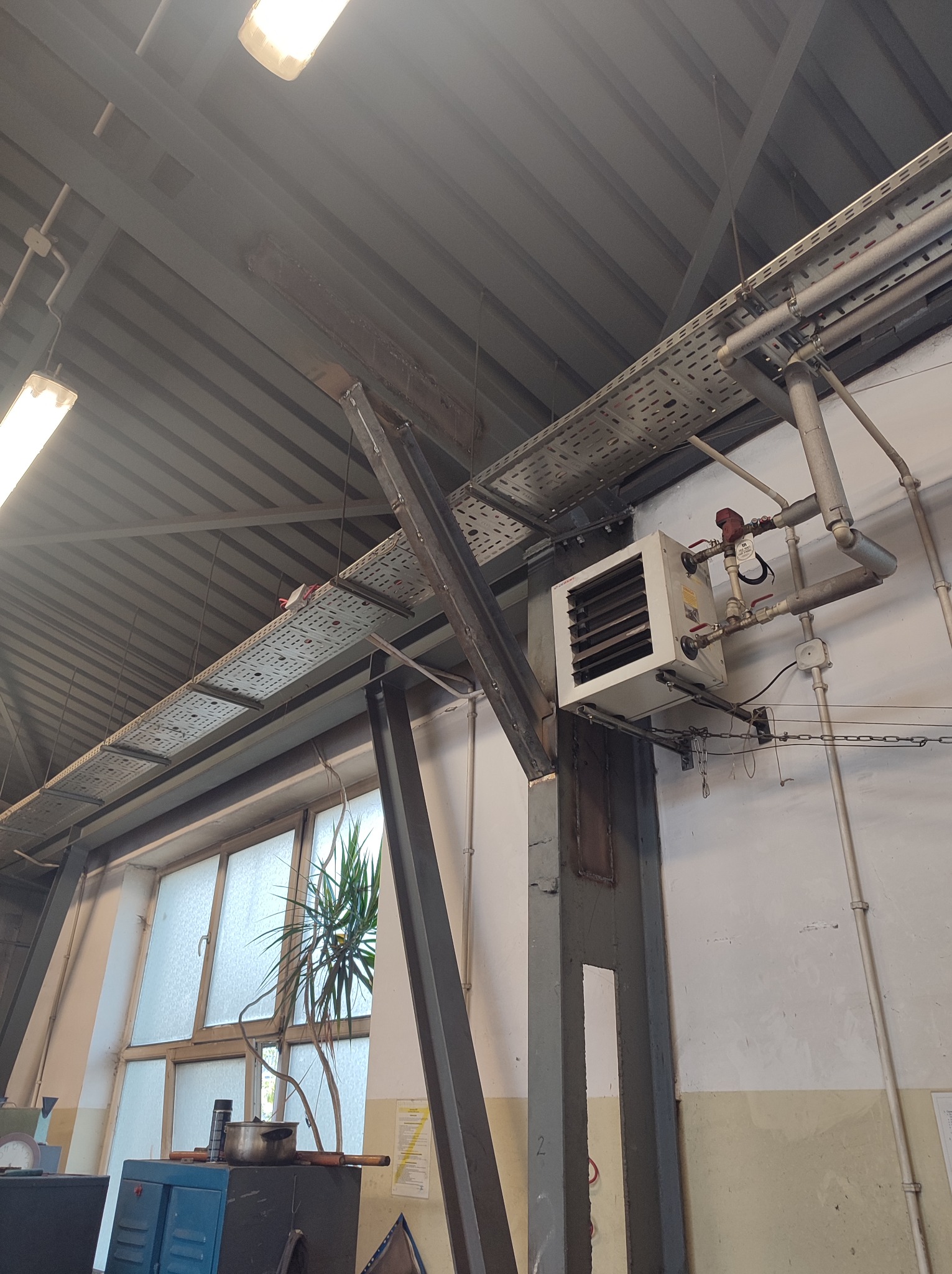 Wnętrze warsztatu z widoczną konstrukcją stalową, instalacją grzewczą pod sufitem, kratkami wentylacyjnymi i fragmentem okna z rośliną doniczkową.