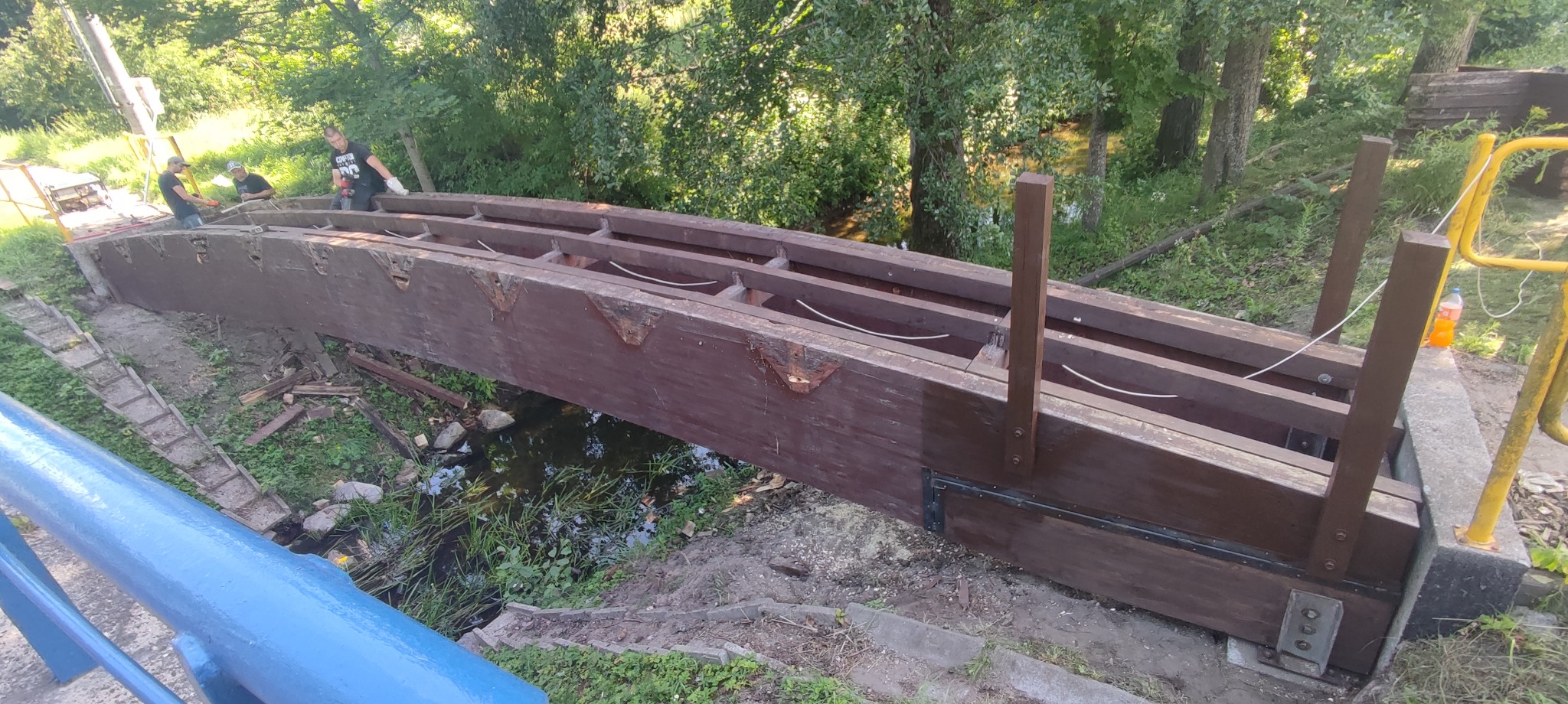 Drewniany most w trakcie budowy nad strumieniem, widoczni robotnicy przy pracy, elementy konstrukcyjne z brązowego drewna, stalowe wzmocnienia, tło z zieleni drzew.