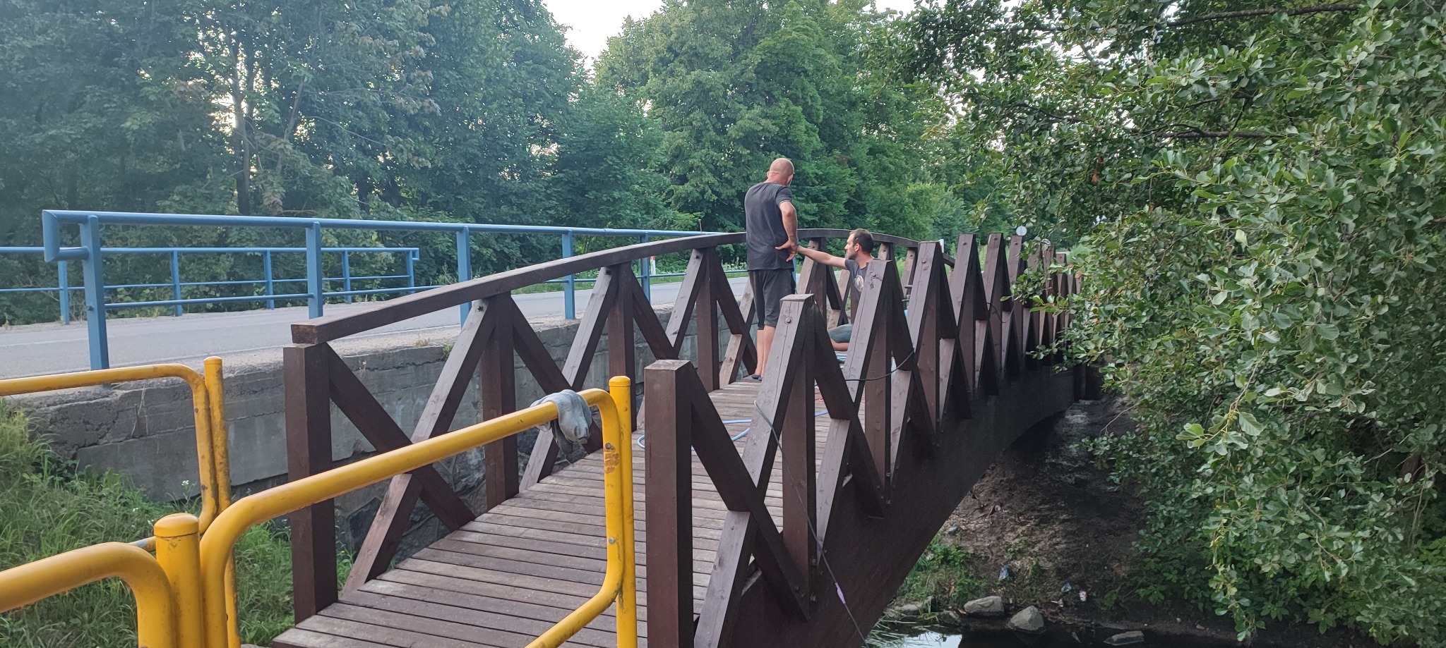 Dwaj mężczyźni rozmawiają na drewnianym moście z żółtymi i niebieskimi elementami, otoczonym zielenią drzew i krzewów.