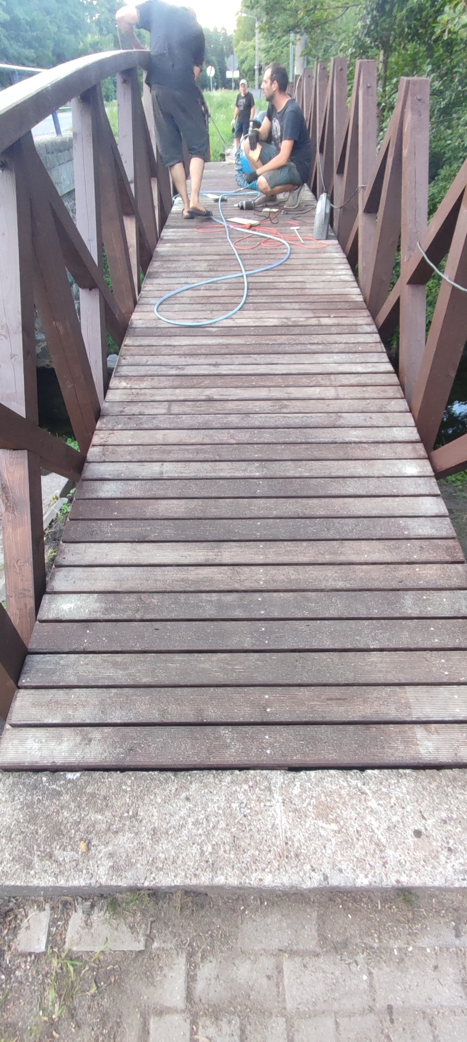 Renowacja drewnianego mostku: pracownicy podczas naprawy desek pomostowych, widoczne narzędzia i przewody pneumatyczne.