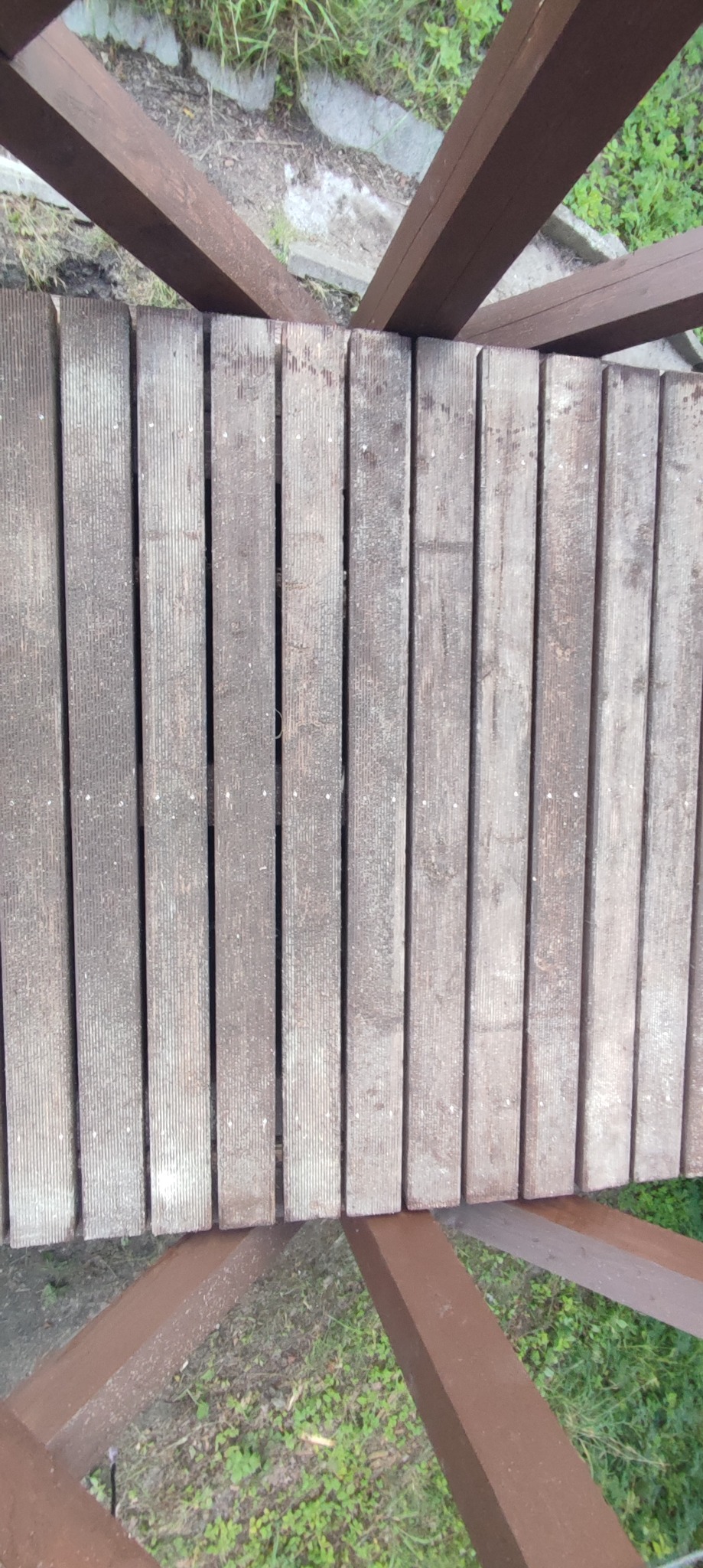 Drewniana konstrukcja schodów zewnętrznych z brązowymi belkami nośnymi, widok z góry na szare, ryflowane deski podestu.