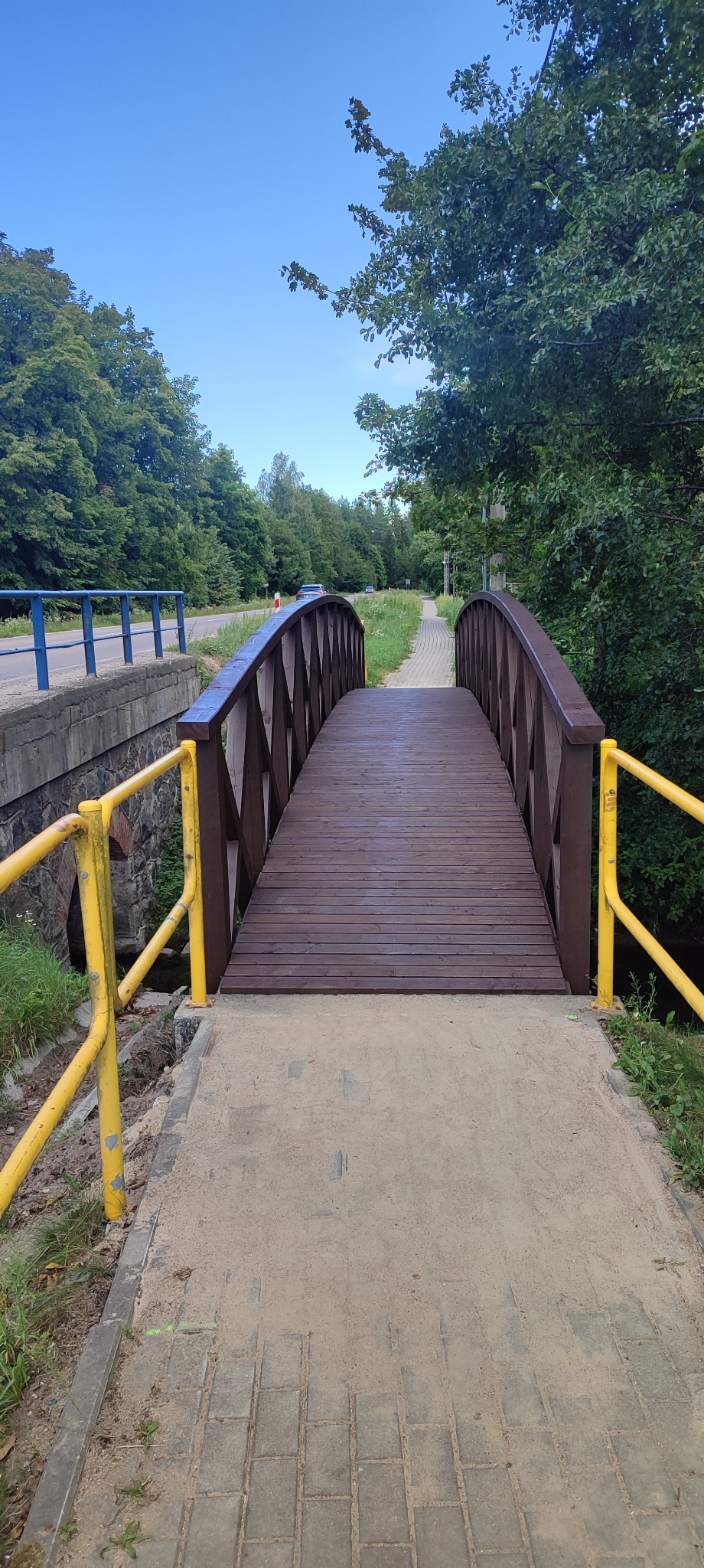 Drewniany mostek łukowy z brązowymi barierkami i żółtymi poręczami, prowadzący nad strumieniem w otoczeniu zieleni, z widoczną drogą w tle.