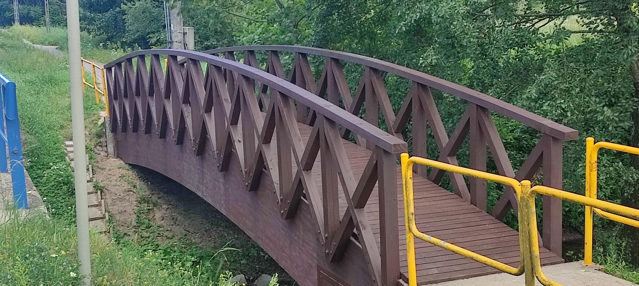Drewniany mostek z brązowymi balustradami i kratownicą, z żółtą barierką po prawej stronie, otoczony zielenią i drzewami.