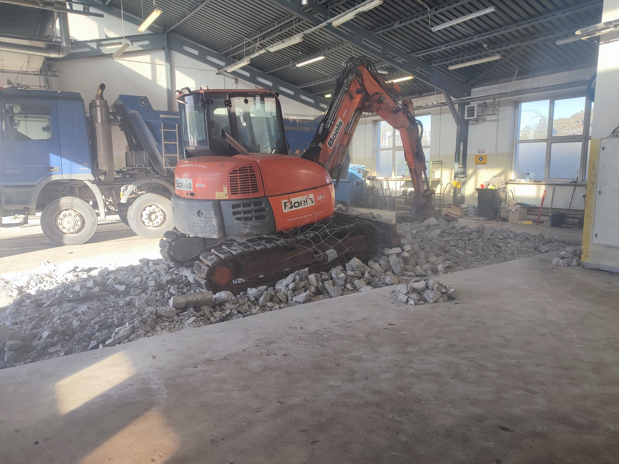 Pomarańczowa minikoparka na gąsienicach kruszy betonowy gruz wewnątrz hali, obok stoi niebieska ciężarówka, widoczne elementy konstrukcji dachu i oświetlenie.