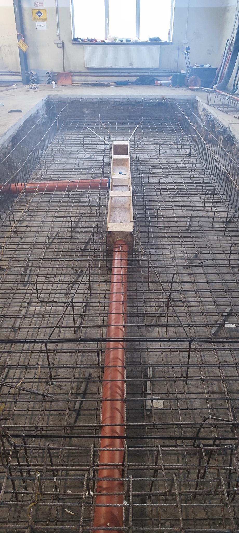 Siatka zbrojeniowa fundamentów z pomarańczową rurą kanalizacyjną i drewnianą formą, widok z góry w wykopie.