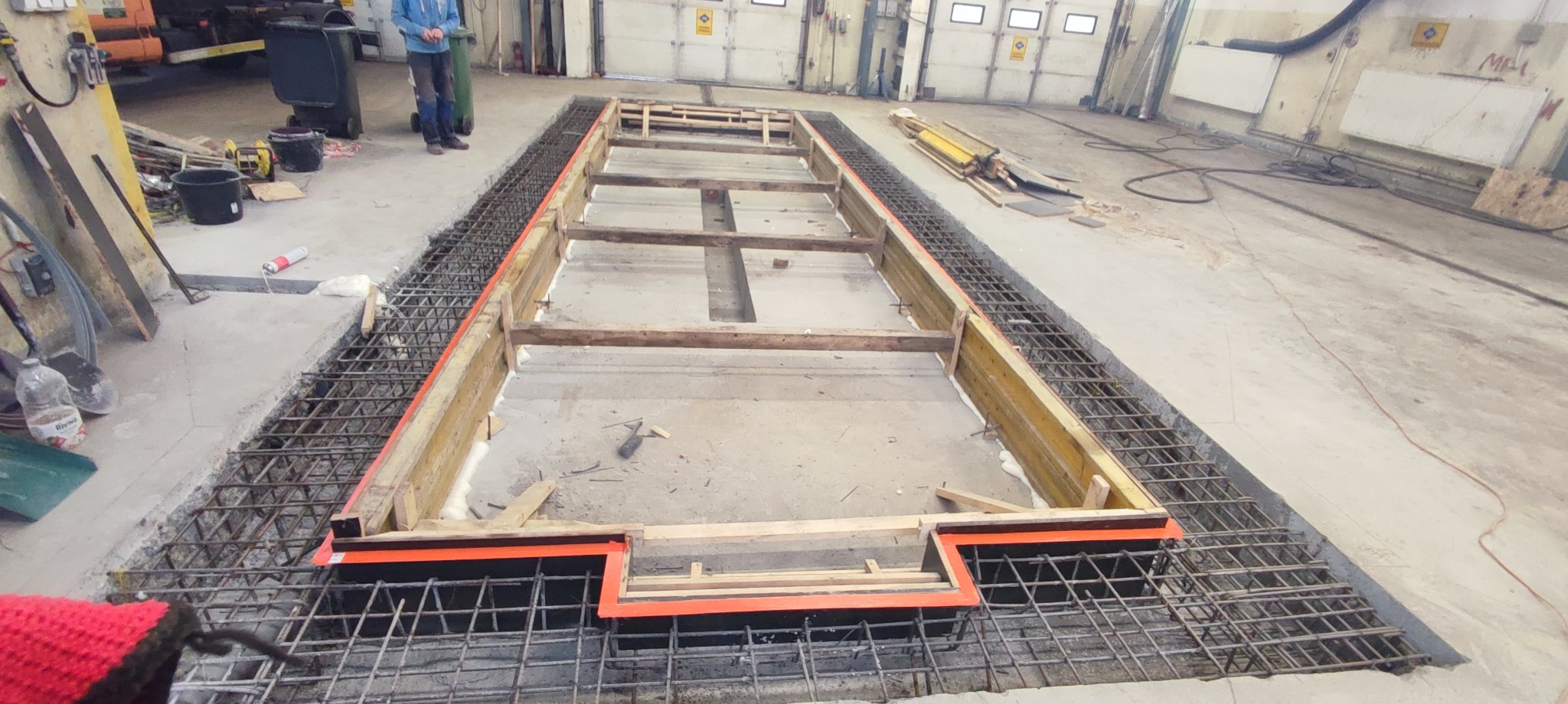 Przygotowanie do wylewki fundamentu w hali: szalunek z drewnianych desek uszczelniony pianką montażową, zbrojenie z siatki stalowej, widoczny fragment podłogi hali i osoba w tle.