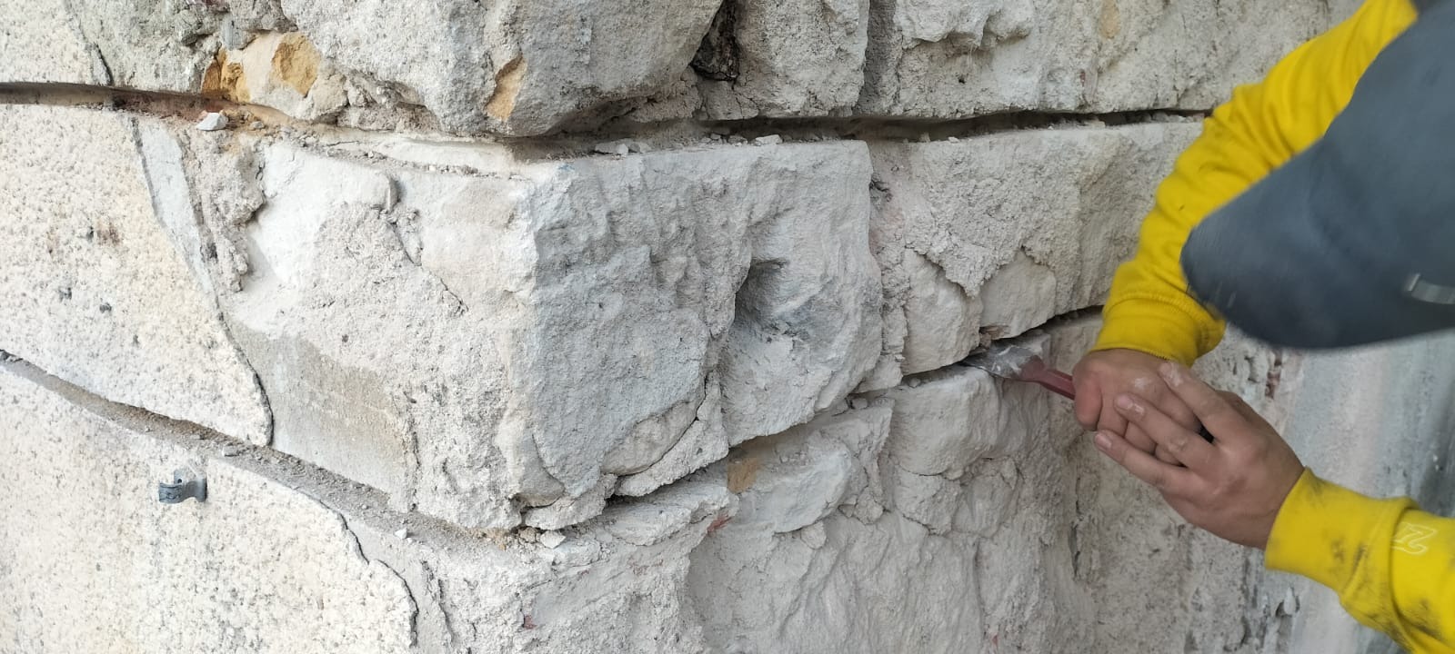 Renowacja starej kamiennej ściany: pracownik w żółtej bluzie używa dłuta do usuwania zaprawy między blokami kamiennymi z widocznym otworem, fragment ściany z metalowym elementem mocującym.