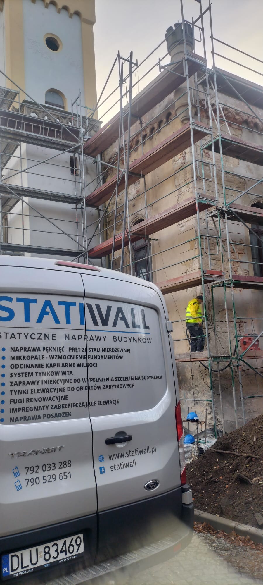 Pracownik w żółtej kamizelce na rusztowaniu podczas renowacji elewacji zabytkowego budynku, z tyłu częściowo widoczny samochód dostawczy firmy Statiwall z reklamą usług naprawy budynków.