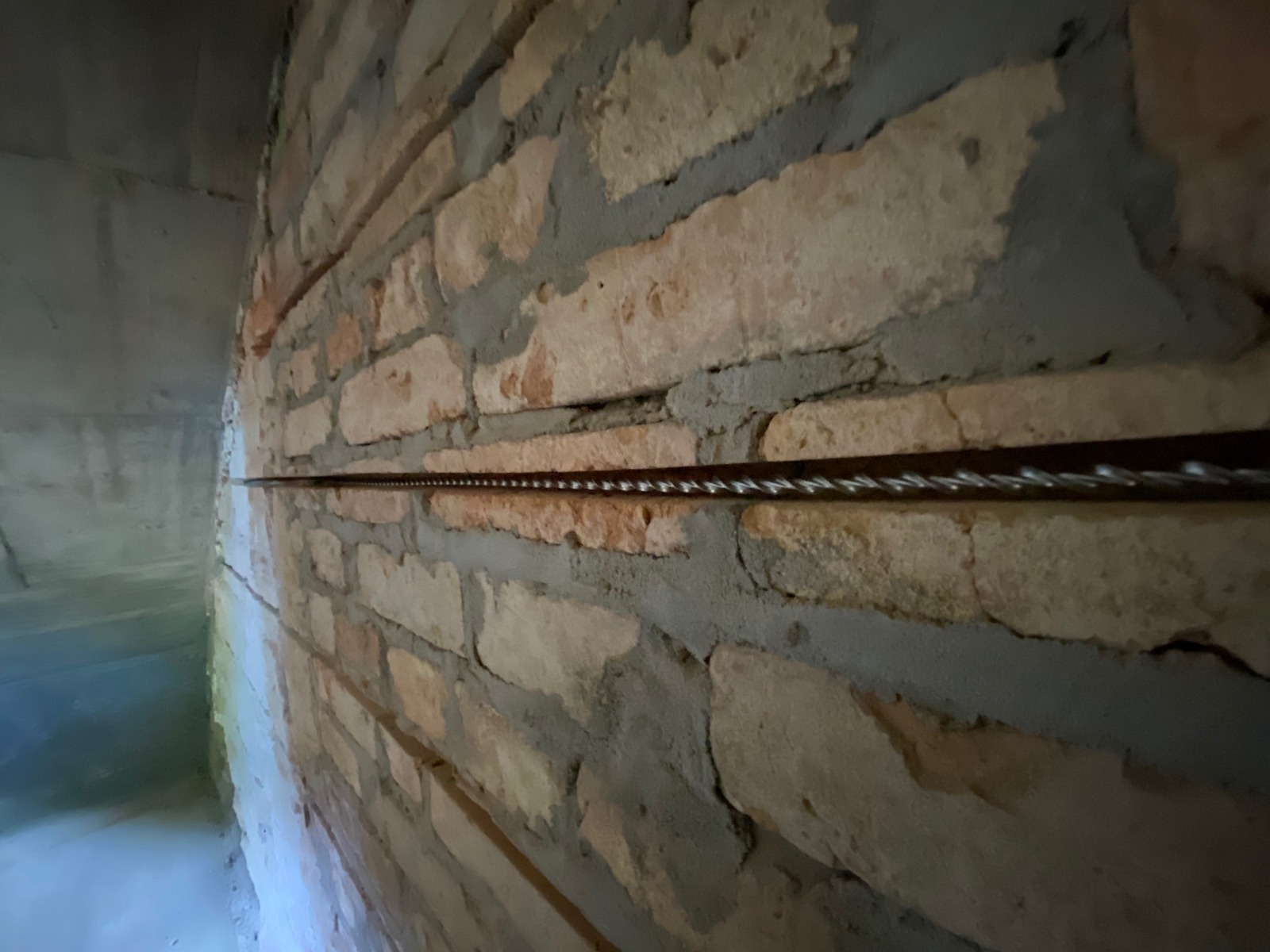 Murowana ściana z cegły z poziomym drutem wzmacniającym w spoinie, widoczne surowe wykończenie i szary beton.