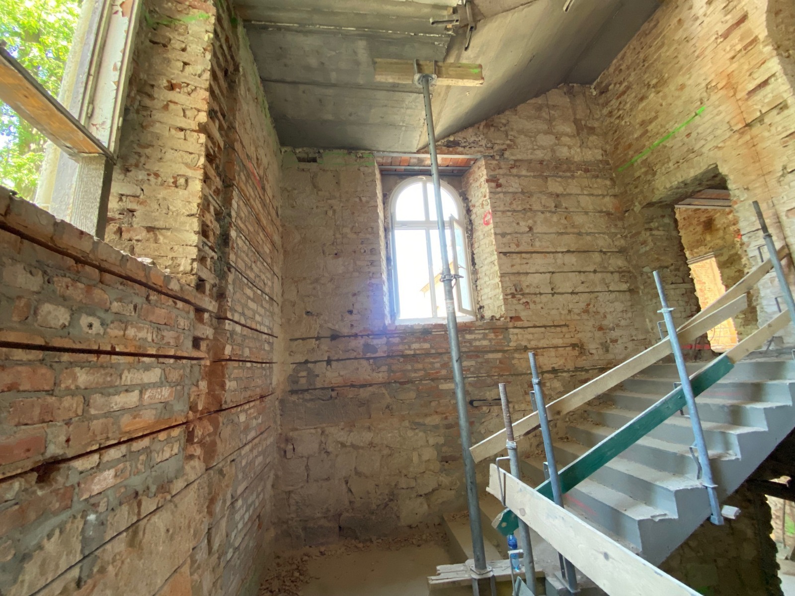 Wnętrze kamienicy w trakcie remontu, widoczne ceglane ściany z poziomymi bruzdami, podpory stropu i betonowe schody w budowie.