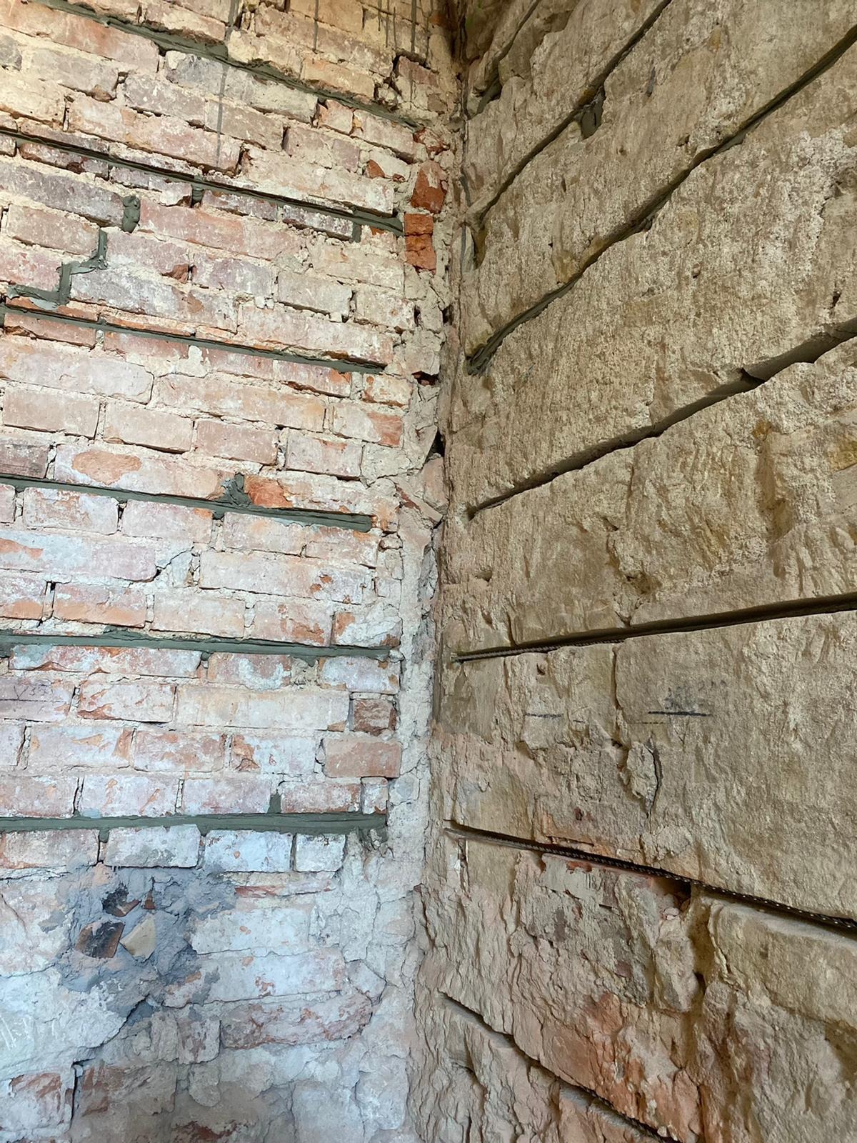 Narożnik ściany z cegły i kamienia z widocznymi poziomymi szczelinami wypełnionymi zaprawą, prawdopodobnie wzmocnienie konstrukcji.