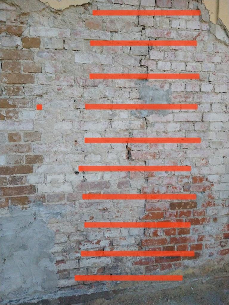 Stara, ceglana ściana z pionowym pęknięciem, zaznaczonym pomarańczowymi liniami w celu monitorowania postępu pękania.
