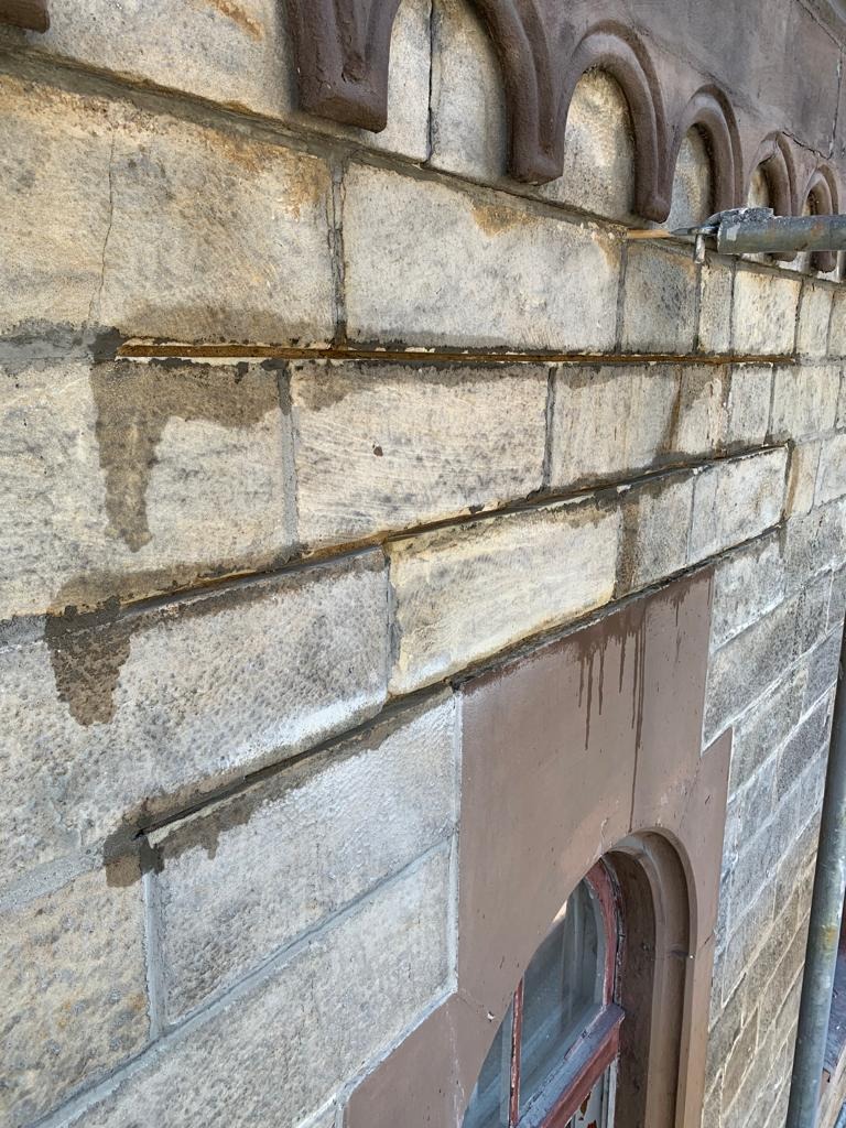 Fragment elewacji kamienicy z widocznymi śladami wilgoci i naprawianymi spoinami między kamieniami; widoczny fragment okna z brązową ramą i rusztowanie.