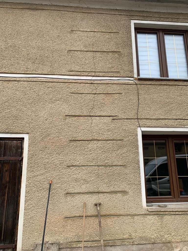 Elewacja budynku z widocznymi pęknięciami i śladami napraw, z drewnianymi oknami i drzwiami, oraz narzędziami opartymi o ścianę.