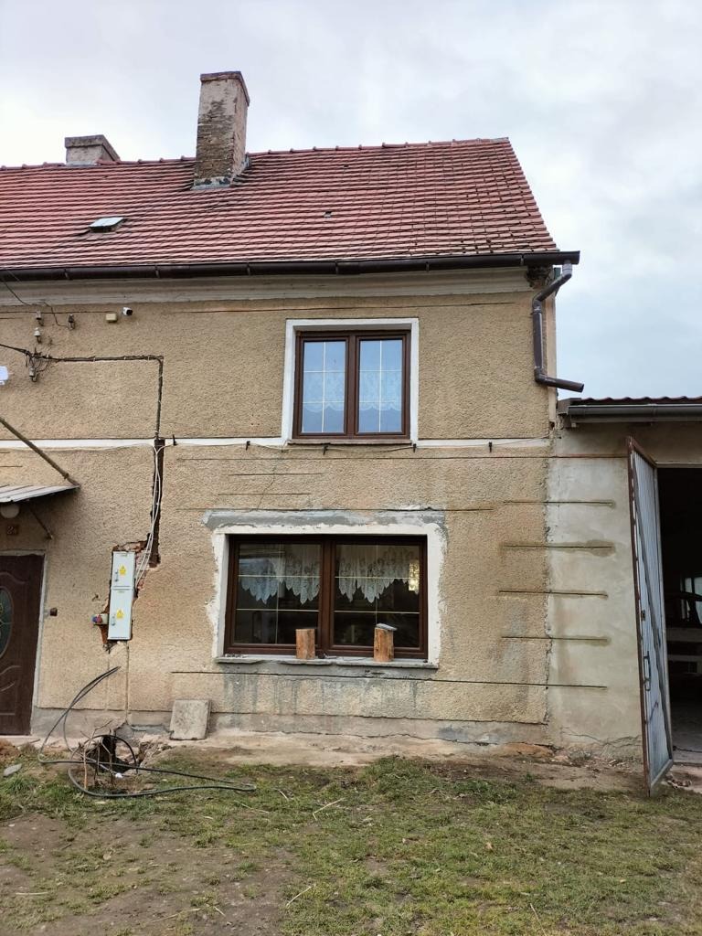 Dwukondygnacyjny dom z oknami zabezpieczonymi drewnianymi belkami, widoczne ślady po remoncie elewacji, otwarte drzwi garażowe, zaniedbany trawnik.