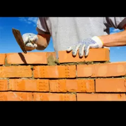 Mężczyzna w rękawicach roboczych układa czerwoną cegłę, używając kielni do nałożenia zaprawy, na tle błękitnego nieba.