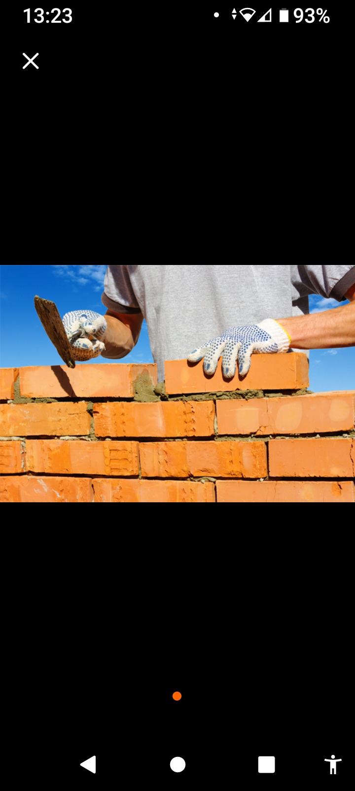 Mężczyzna w rękawicach roboczych układa czerwoną cegłę, używając kielni do nałożenia zaprawy, na tle błękitnego nieba.